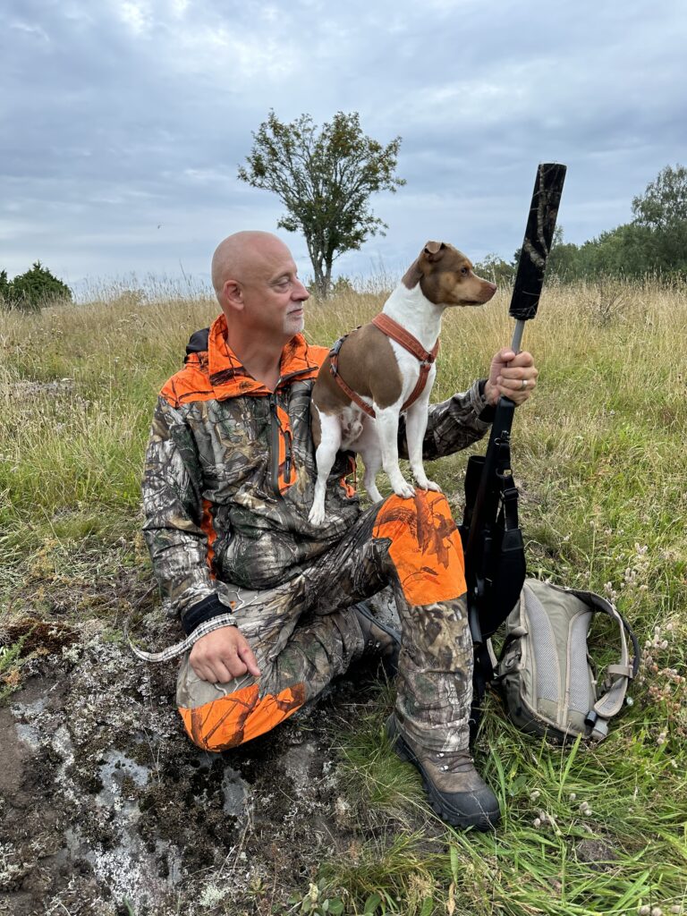 Cazador con ropa naranja y de camuflaje junto a su perro en el campo, con arma y equipo de protección